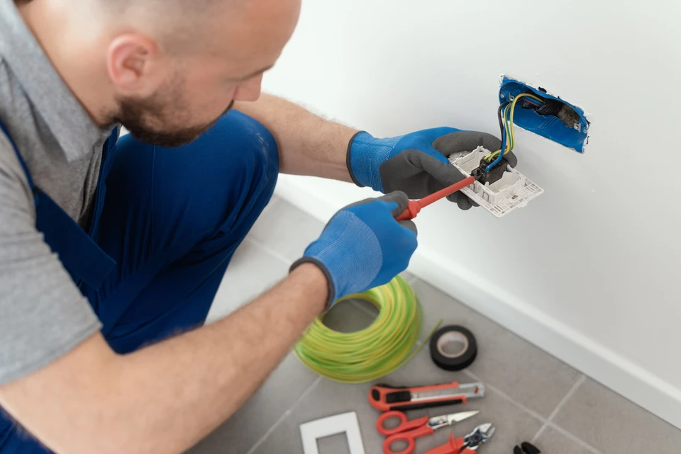 Électricien en combinaison bleue installant une prise électrique murale dans un intérieur résidentiel blanc